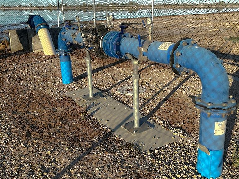 A large blue pipe coming up from the ground, through a valve and back into the ground. Shallow lake in background.